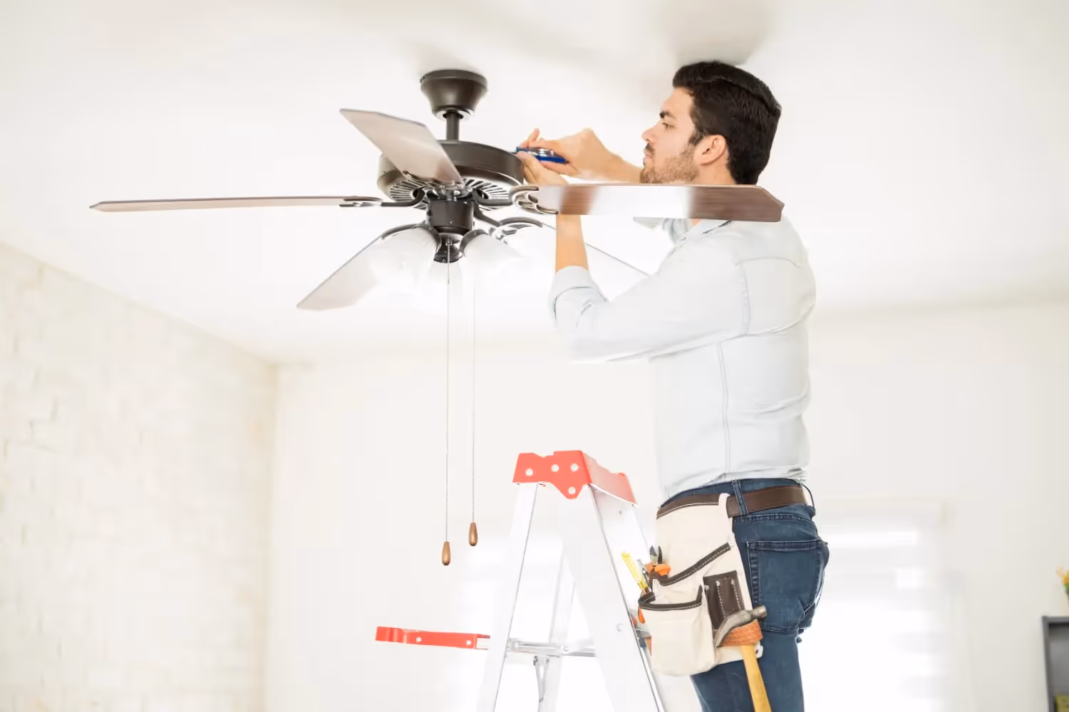 A technician standing on a ladder and wearing a tool belt uses a screwdriver to install a dark ceiling fan.