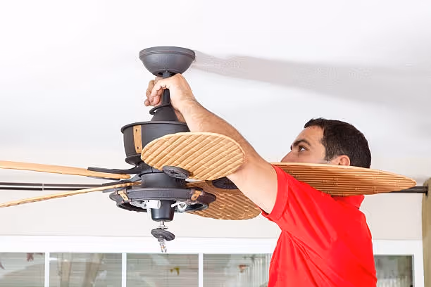 A man in a red shirt installs a ceiling fan with wooden blades, focusing intently.