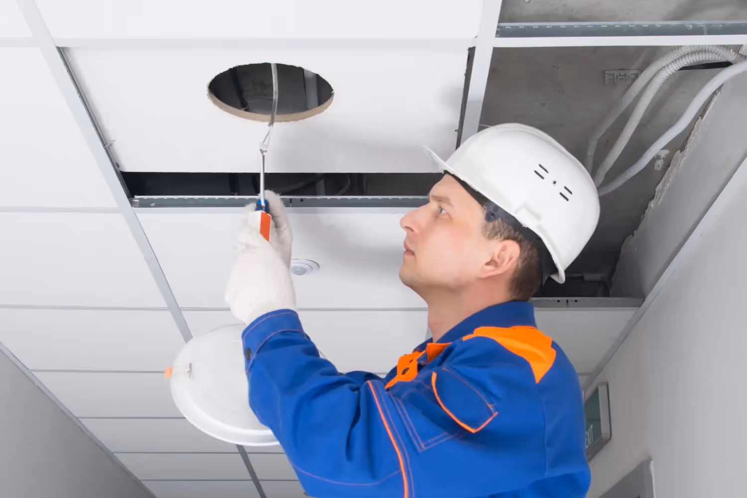 An electrician in a blue uniform and white hard hat installs a circular light fixture into a suspended ceiling. He is reaching into the tile opening to manage the wiring.