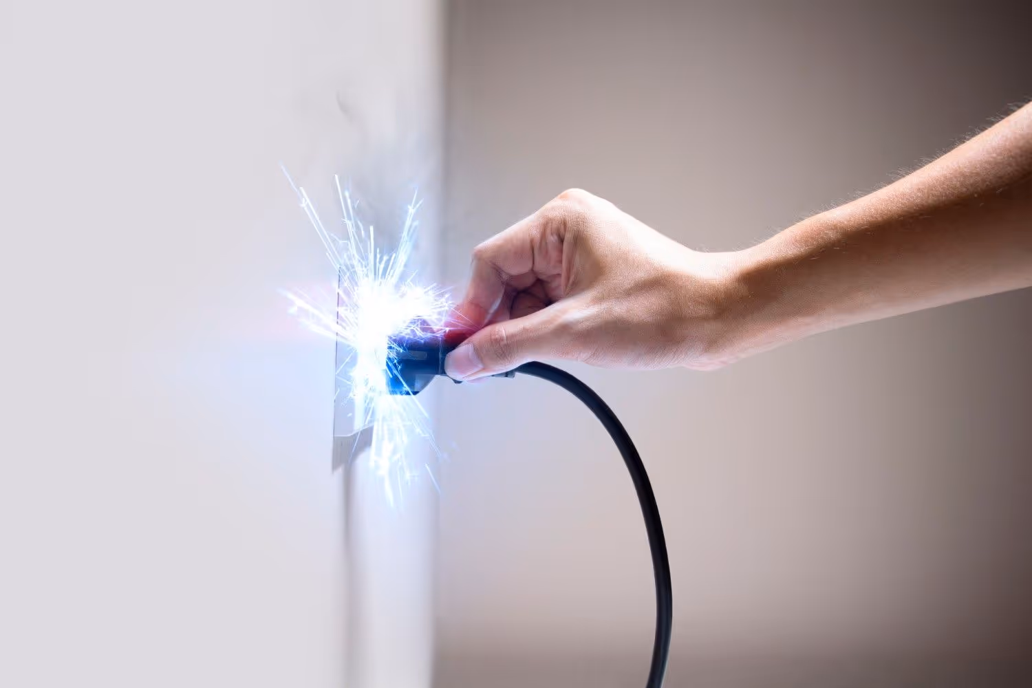 Bright blue electrical sparks erupt from a wall outlet as a person attempts to plug in a black power cord. Wisps of smoke rise from the socket against a plain, light-colored wall.