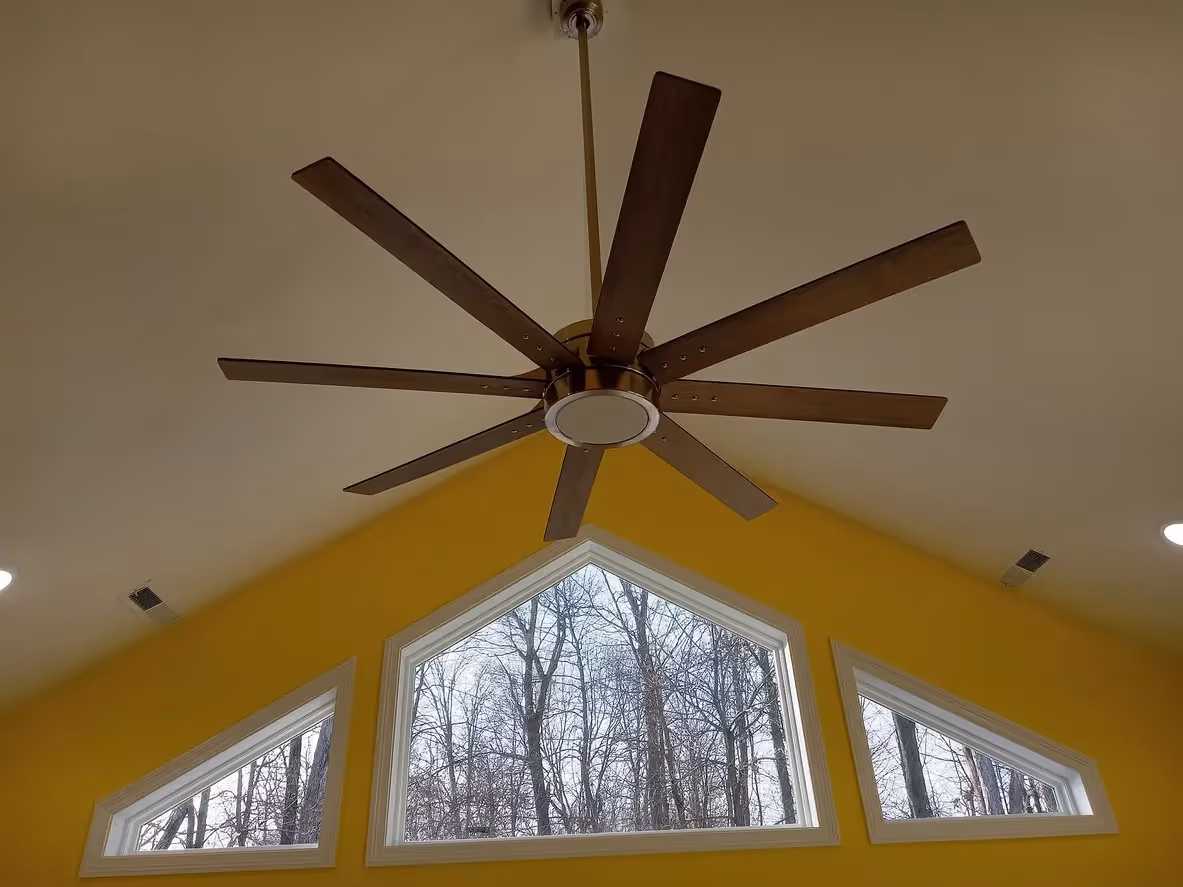 Low-angle view of a large, eight-blade wooden ceiling fan with a central light fixture, mounted on a high white ceiling above a yellow wall with three geometric windows.