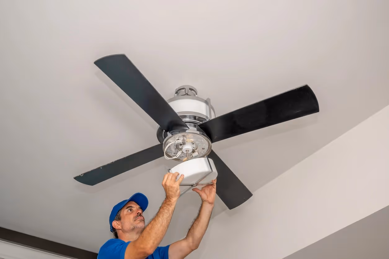 A technician in a blue cap and t-shirt reaches upward to install a white frosted glass light fixture onto a four-blade black ceiling fan mounted on a plain white ceiling.