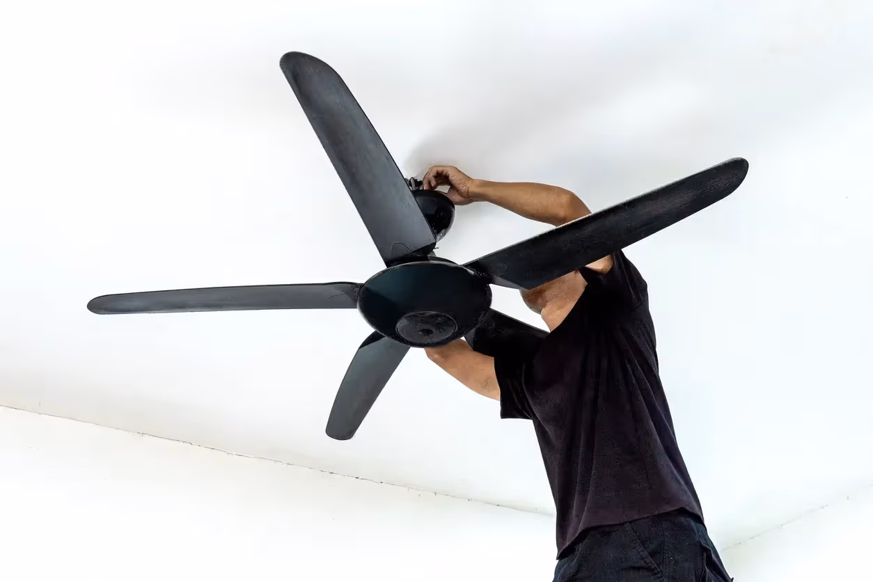 Low-angle view of a man in a black t-shirt reaching upward to install or repair a four-blade black ceiling fan mounted on a plain white ceiling.