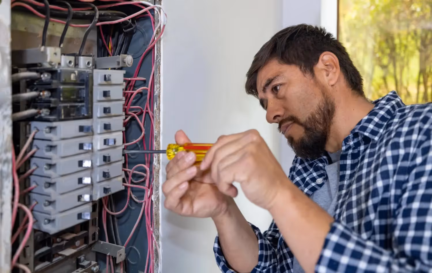 A male electrician in a plaid shirt uses a yellow-handled screwdriver on an electrical wiring panel.