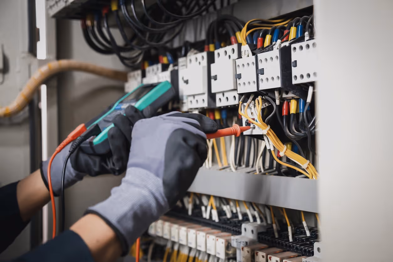 Electrician wearing gloves uses a multimeter to test wires in a complex electrical panel.
