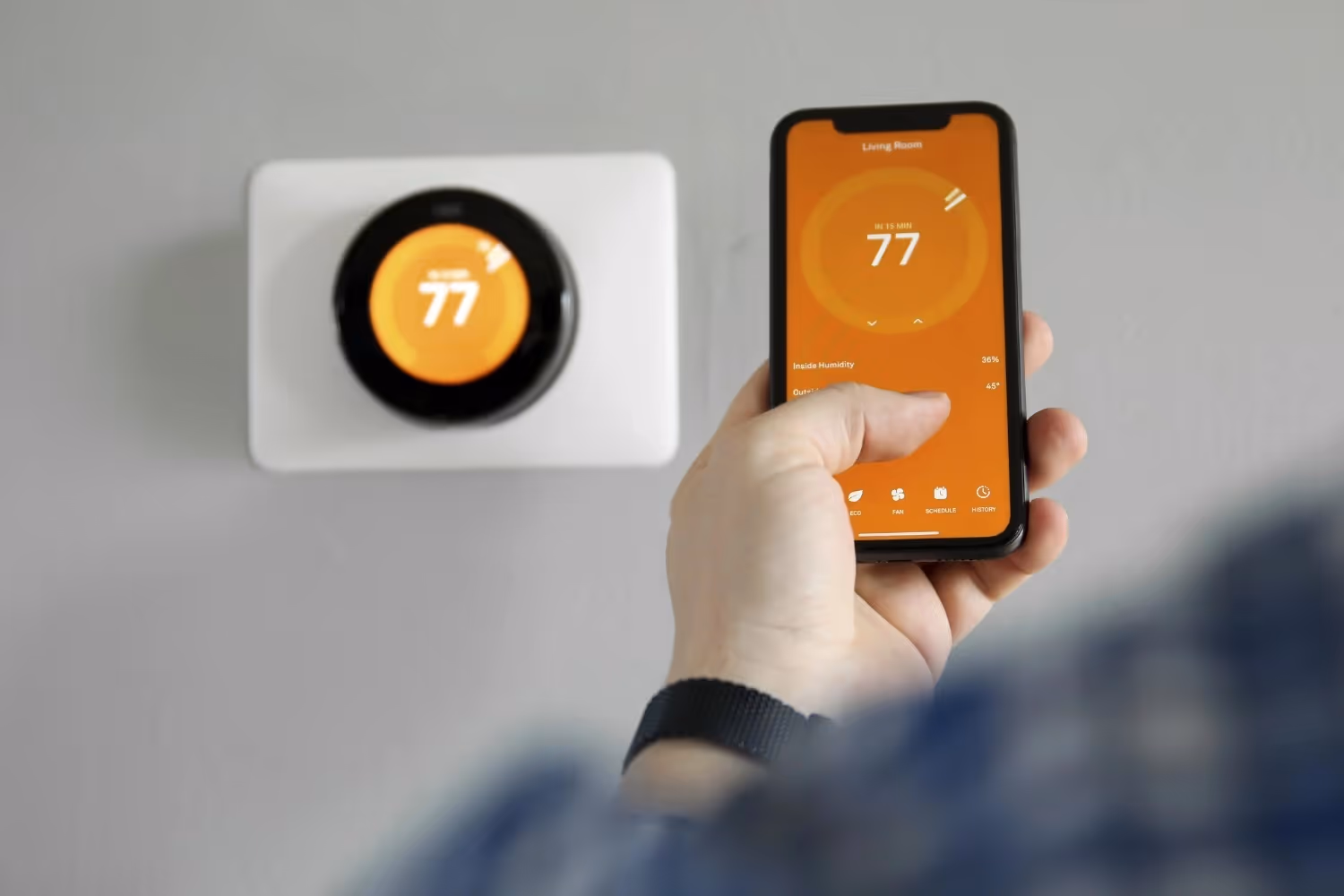 A hand holding a smartphone displaying a thermostat app next to a wall-mounted smart thermostat.