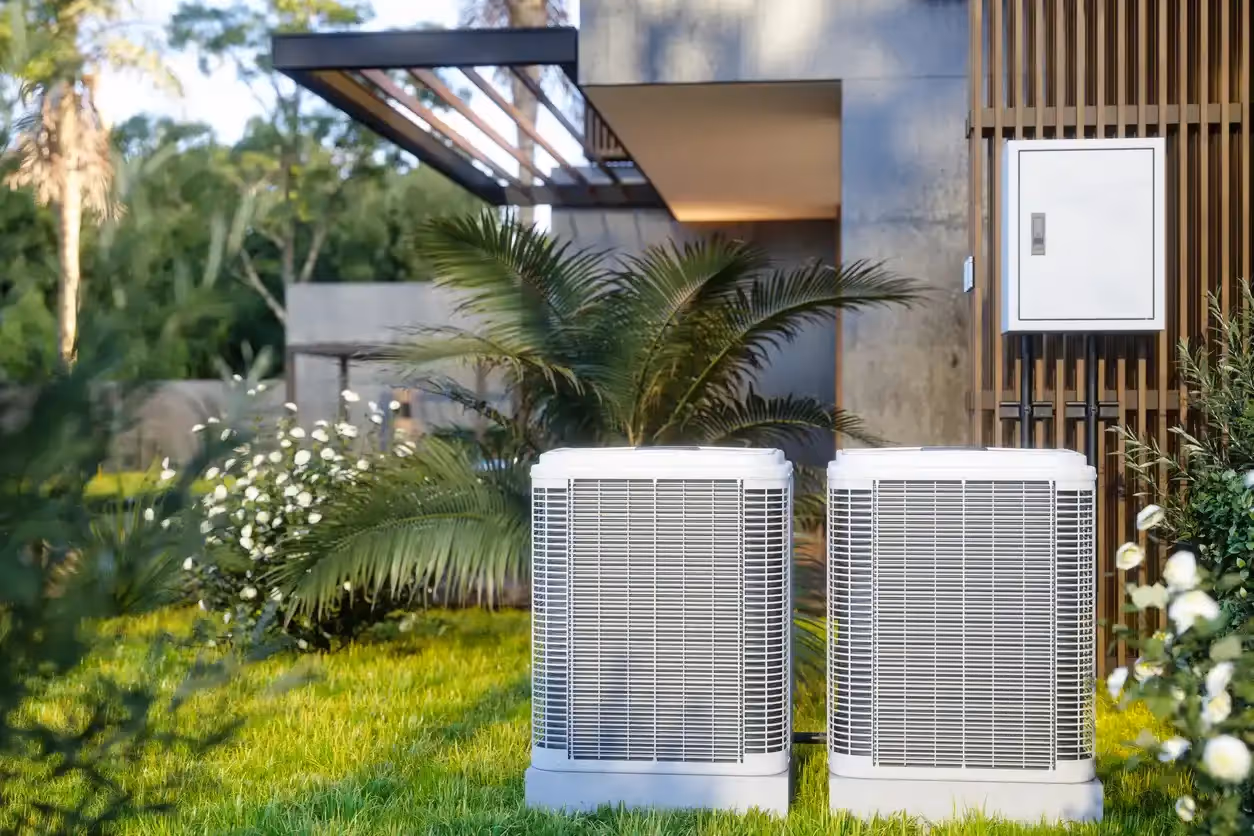 Two identical, modern white heat pump units are installed on a lush green lawn next to a contemporary house with a garden and palm trees.