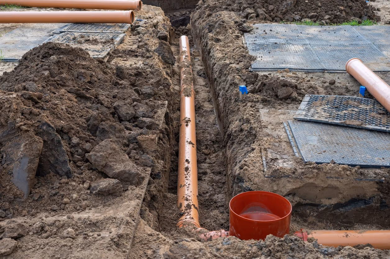 A construction site with exposed dirt shows orange drainage pipes laid in trenches, alongside metal grates. 