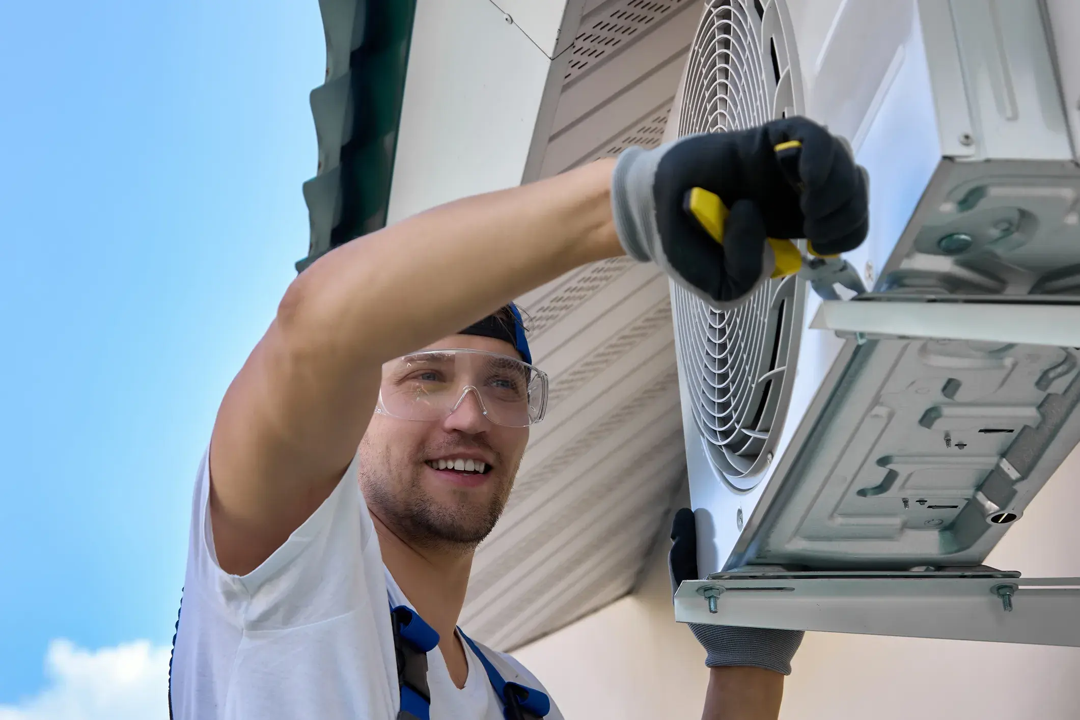 Smiling HVAC technician in safety glasses repairing the fan section of an outdoor AC unit.