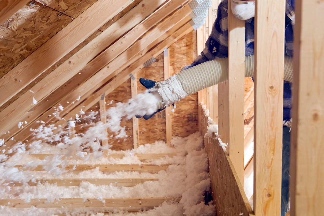 A worker uses a hose to blow white insulation into an attic with wooden beams.