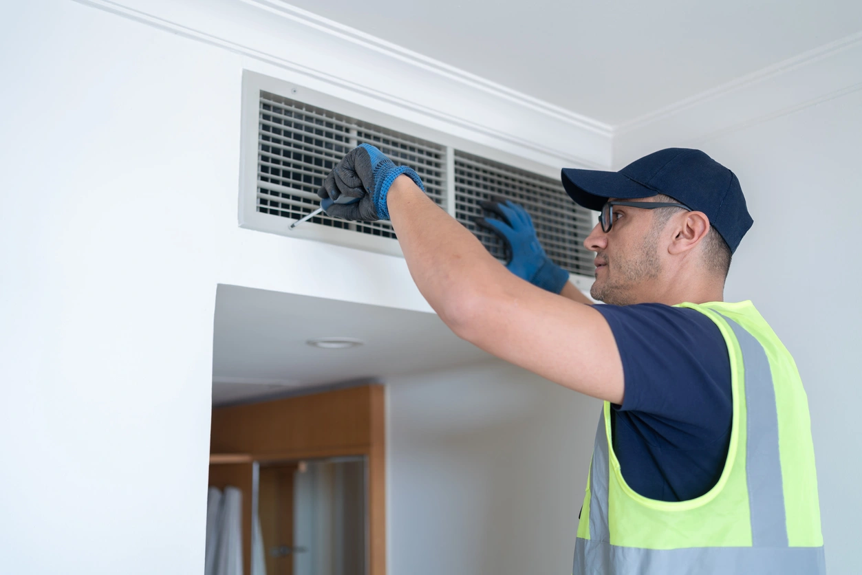 A technician in a cap and safety vest inspects an air vent on a white ceiling. 