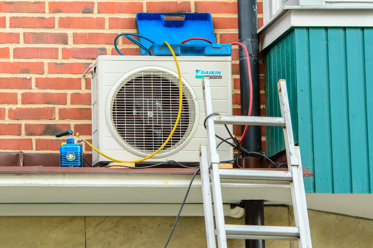 Outdoor air conditioning unit on a brick wall, with hoses and a blue toolbox on top.