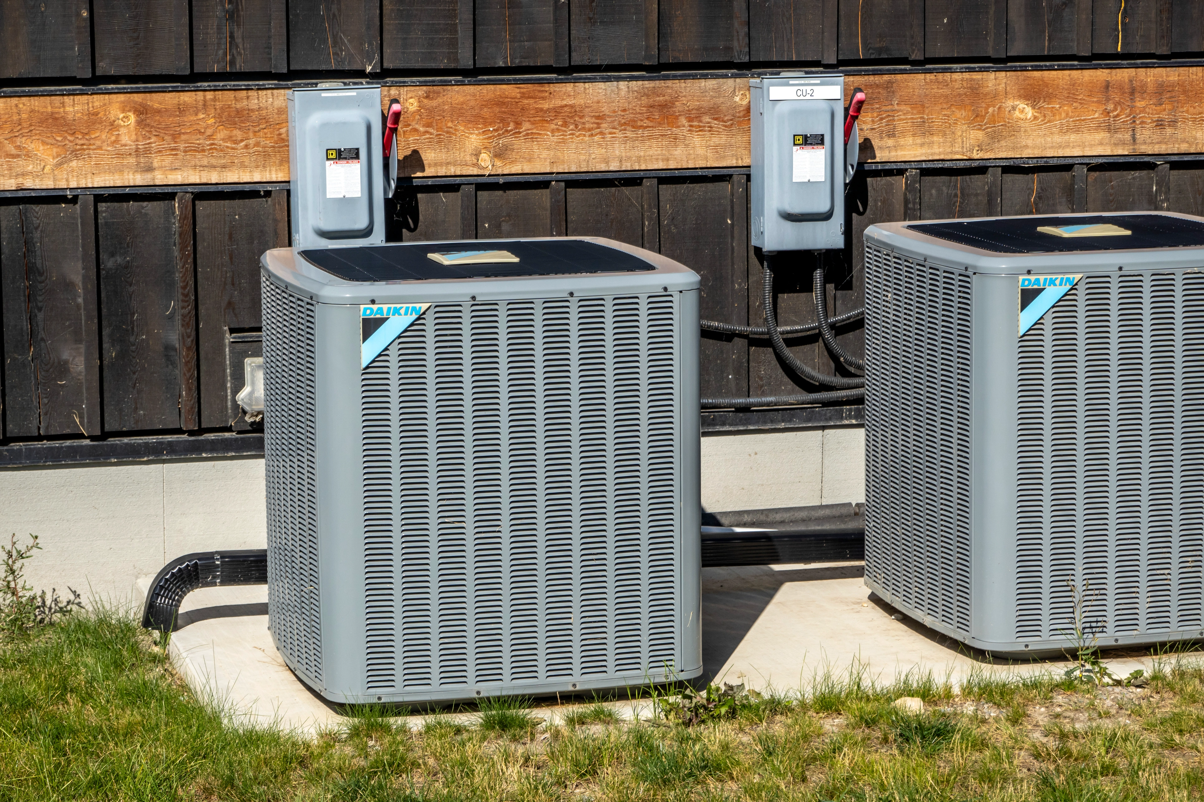 Two gray outdoor air conditioning units are installed against a wooden wall on a sunny day, surrounded by grass. 