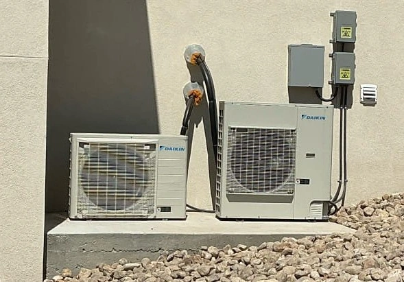 Outdoor air conditioning units on a concrete platform against a beige wall, surrounded by rocks.