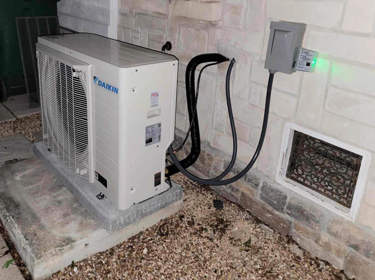 Outdoor air conditioning unit on a concrete pad next to a brick wall. Nearby electrical box with glowing green light.