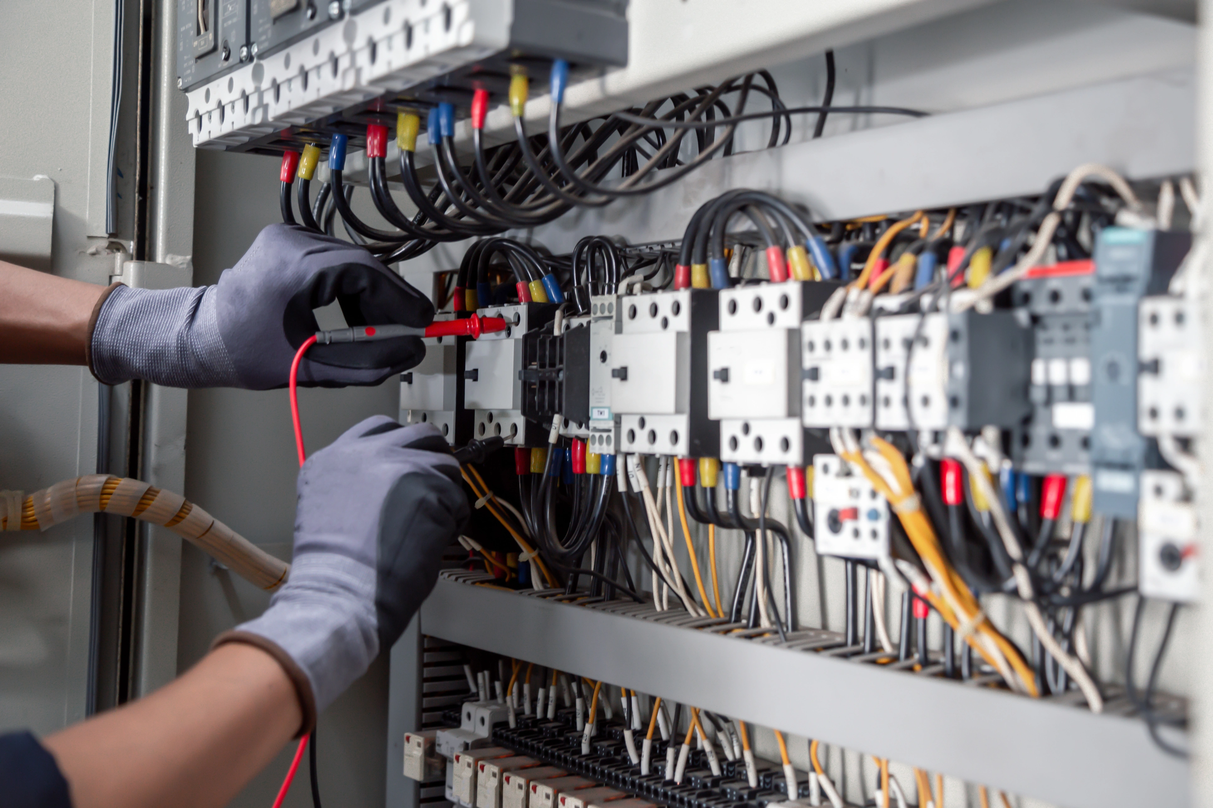 Electrician's gloved hands testing wires in a complex electrical panel. 