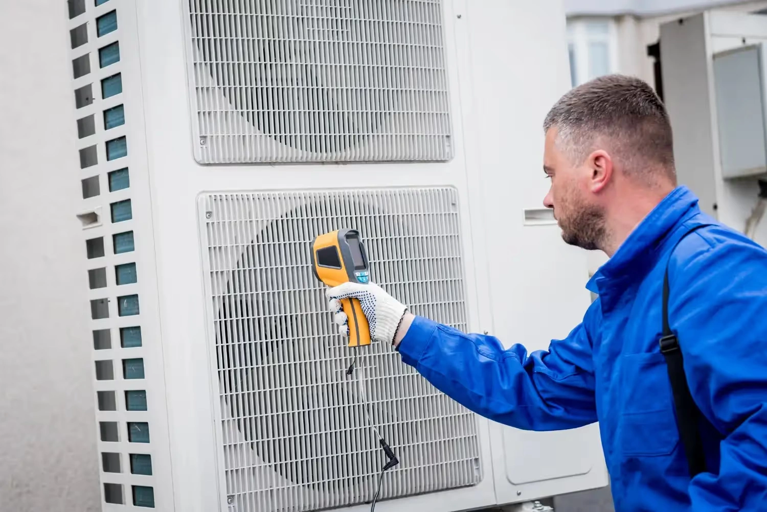 A technician in a blue uniform uses a thermal scanner on a large air conditioning unit.