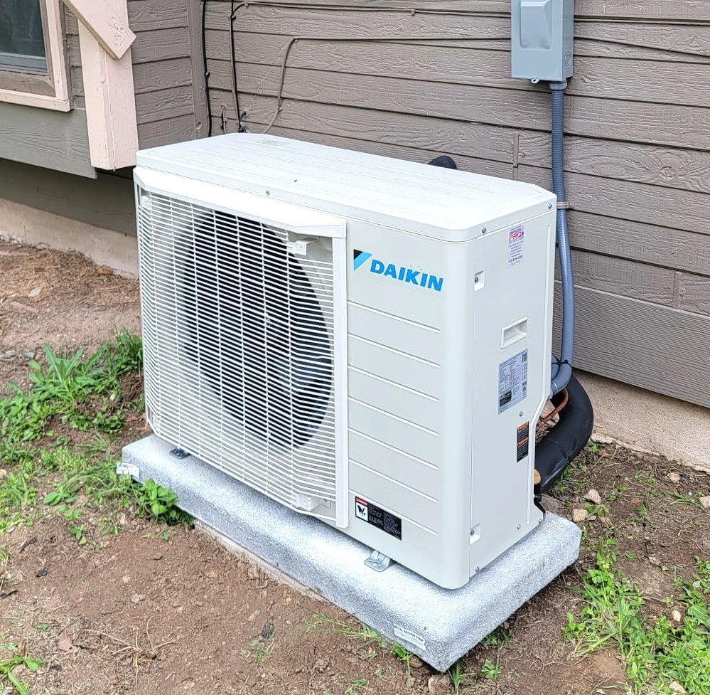 Outdoor Daikin HVAC unit on a concrete base near a wooden house wall, surrounded by grass.