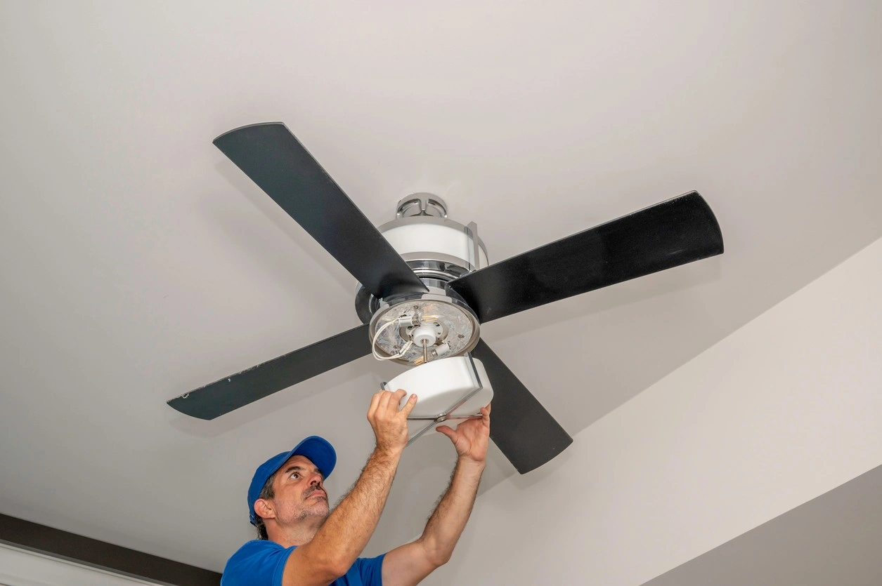 A technician in a blue cap and t-shirt reaches upward to install a white frosted glass light fixture onto a four-blade black ceiling fan mounted on a plain white ceiling.