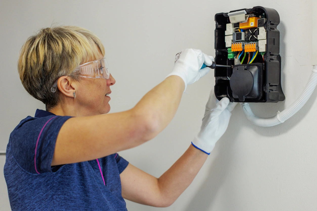 A person in safety glasses and gloves is inspecting electrical wires in an open wall-mounted box.