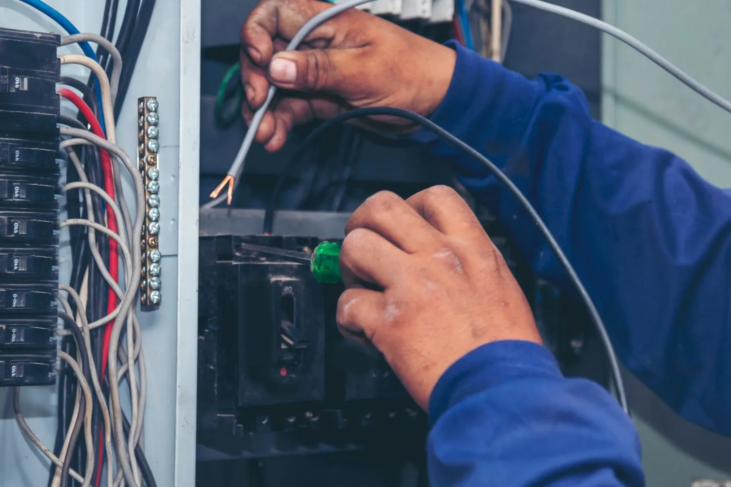 A close-up of a person's hands wearing a blue shirt, using a screwdriver to fix an electrical panel.