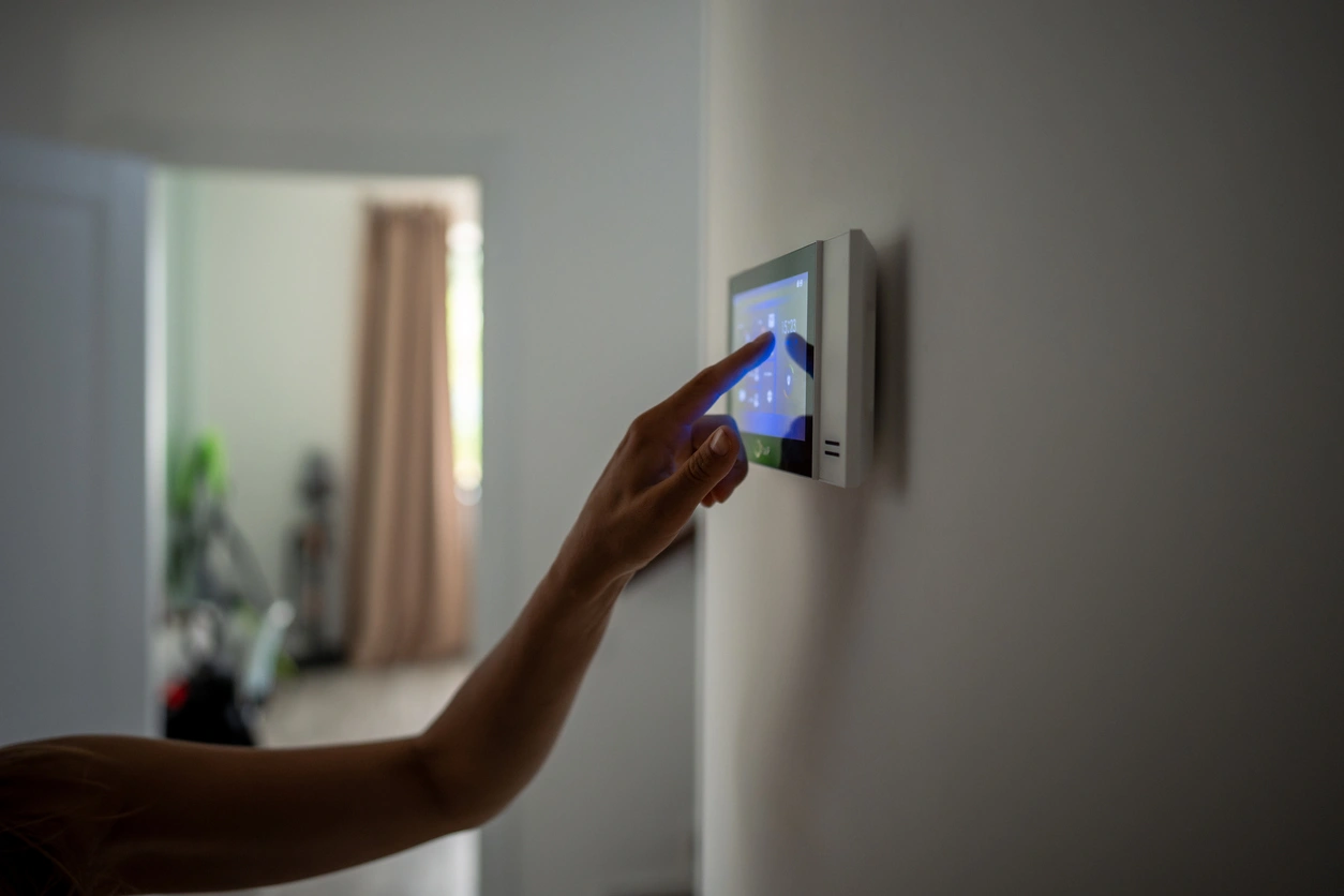 A hand reaches to adjust a digital thermostat on a wall, with soft lighting and a blurred room in the background.