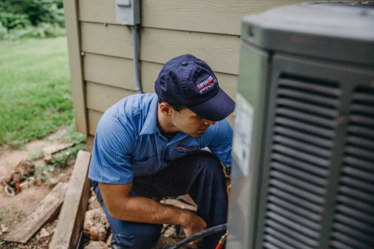 A technician in a blue uniform and cap examines an outdoor air conditioning unit beside a beige house. 