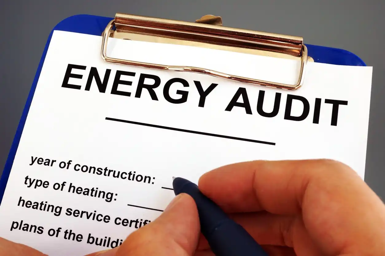 A hand holds a pen, poised to fill out an "Energy Audit" form on a clipboard. 