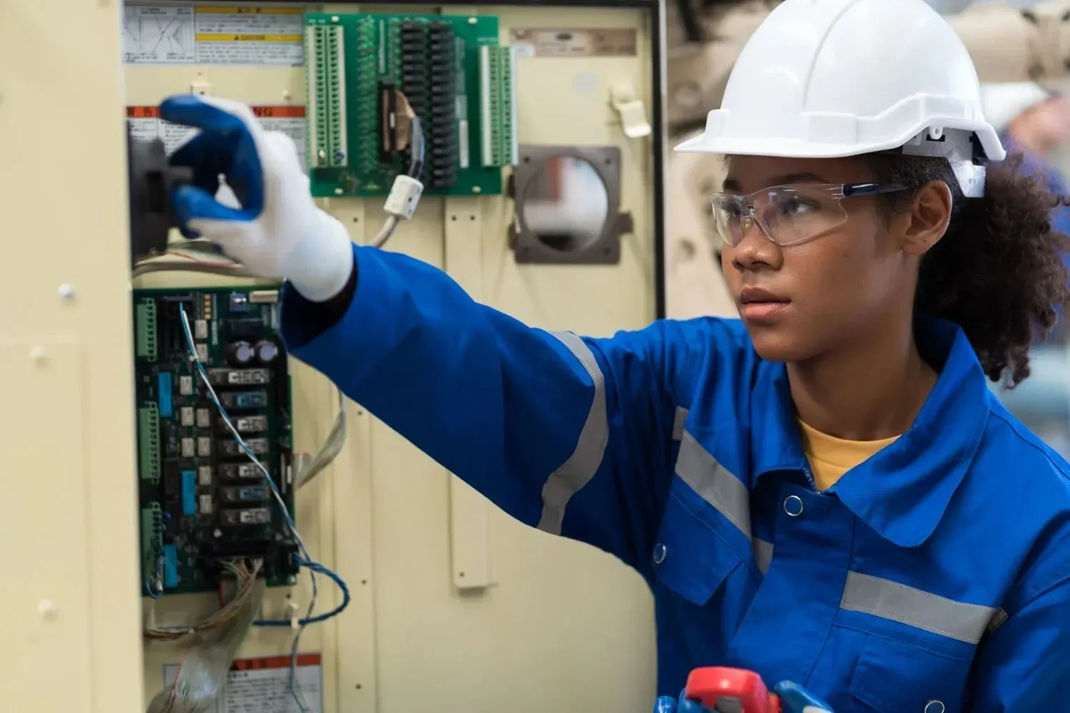 An industrial worker in a hard hat and safety gear performs an inspection of an electrical control panel.