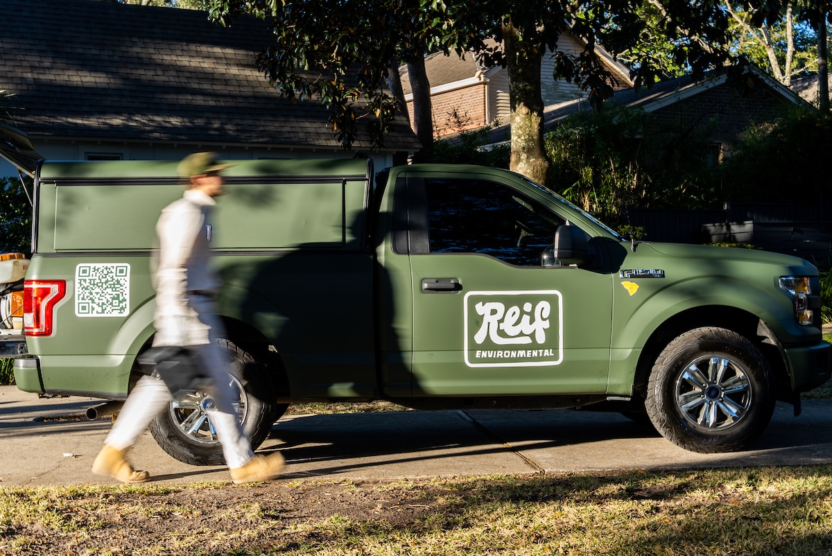 Green pickup truck with Reif Environmental logo parked on a driveway as a person in light clothing and boots walks past.