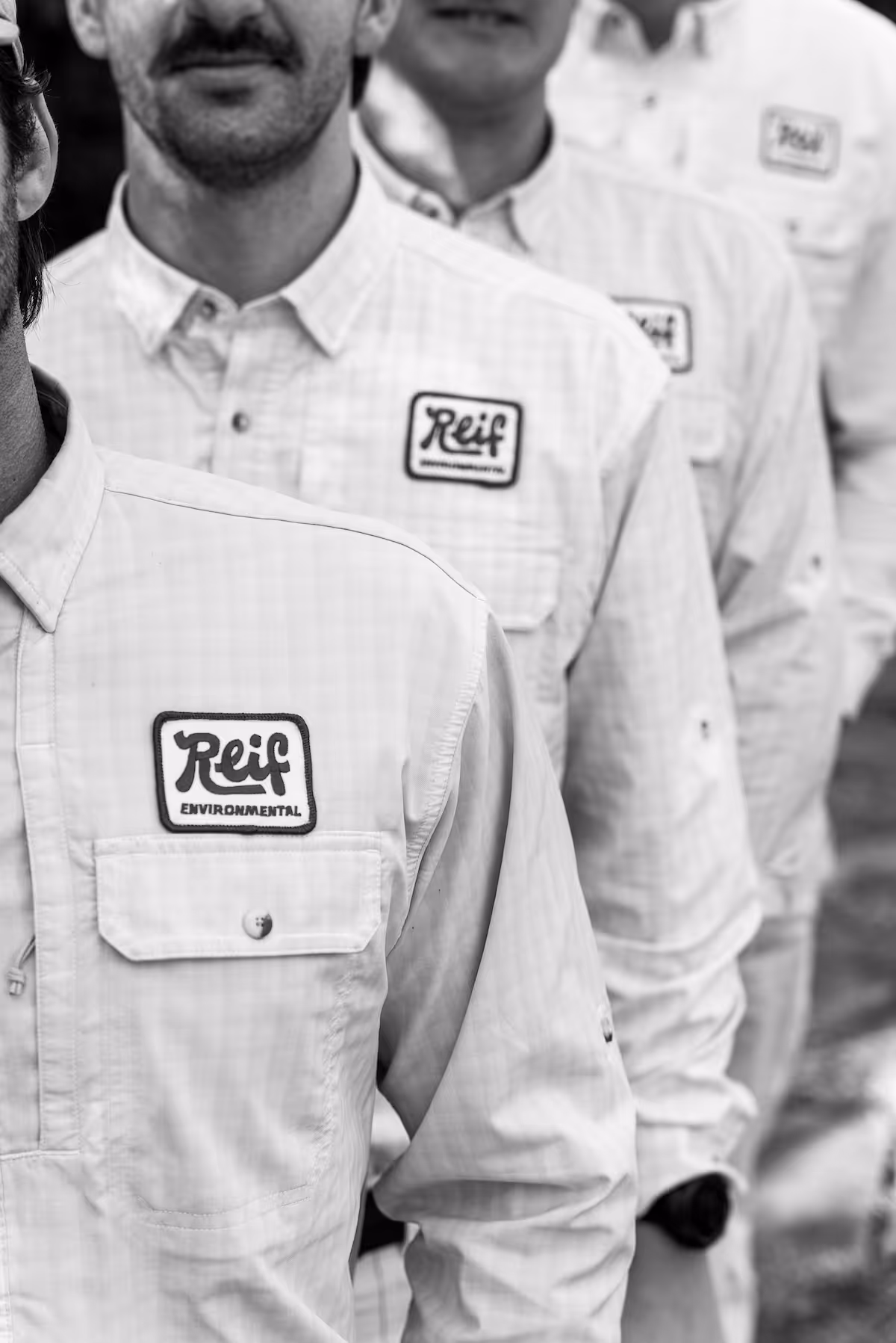 Close-up of three men standing in a row wearing matching light-colored shirts with 'Reif Environmental' patches.