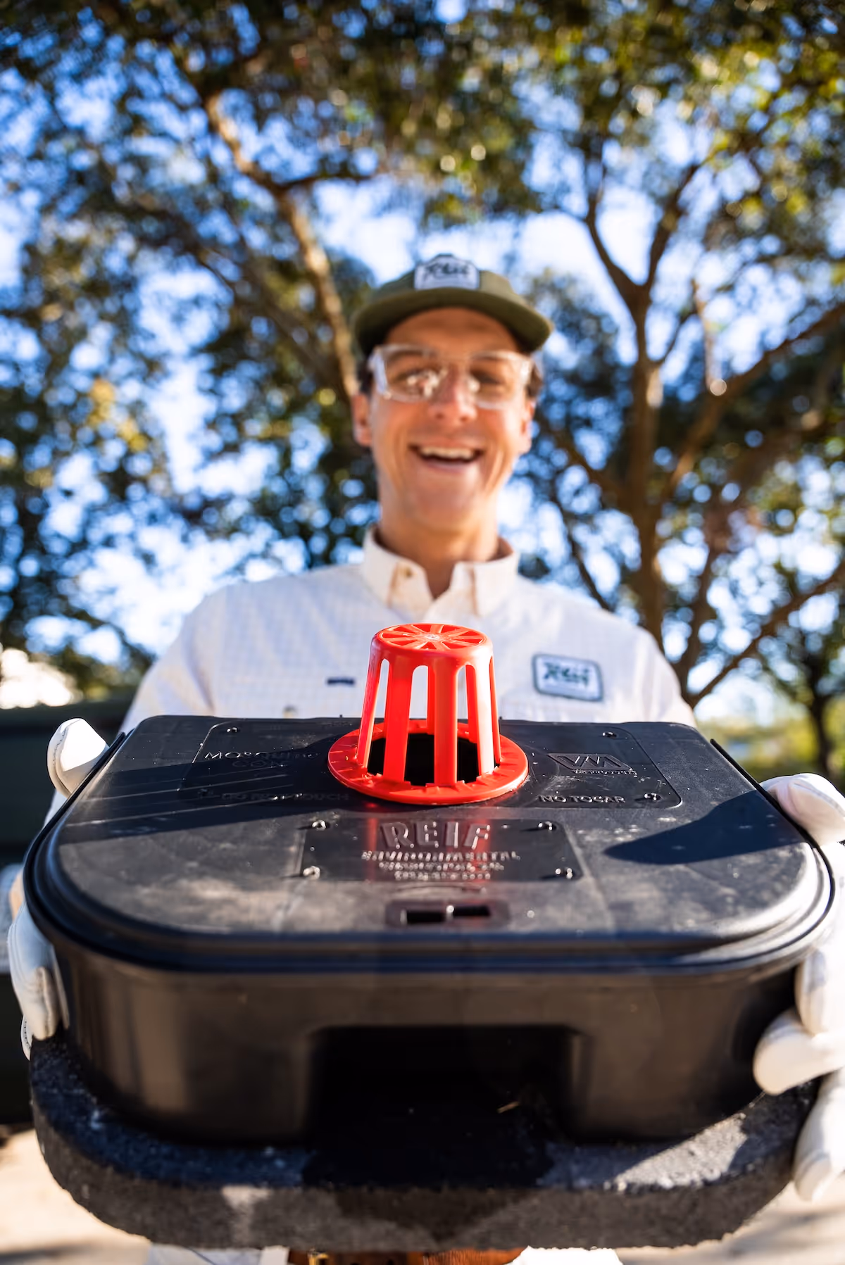 Smiling man in safety glasses and gloves holding a black mosquito control device with a red vent on top outdoors.