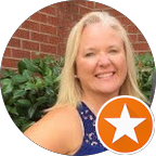 Smiling blonde woman in a blue sleeveless top standing outdoors with greenery and a brick wall in the background.