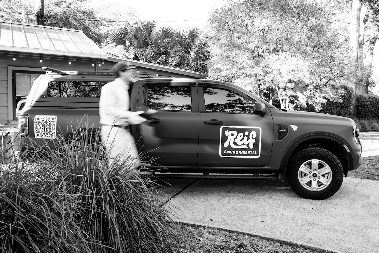 Blurred person walking beside a pickup truck with Reif Environmental logo and a QR code on its side, parked in a driveway with plants and a house in the background.
