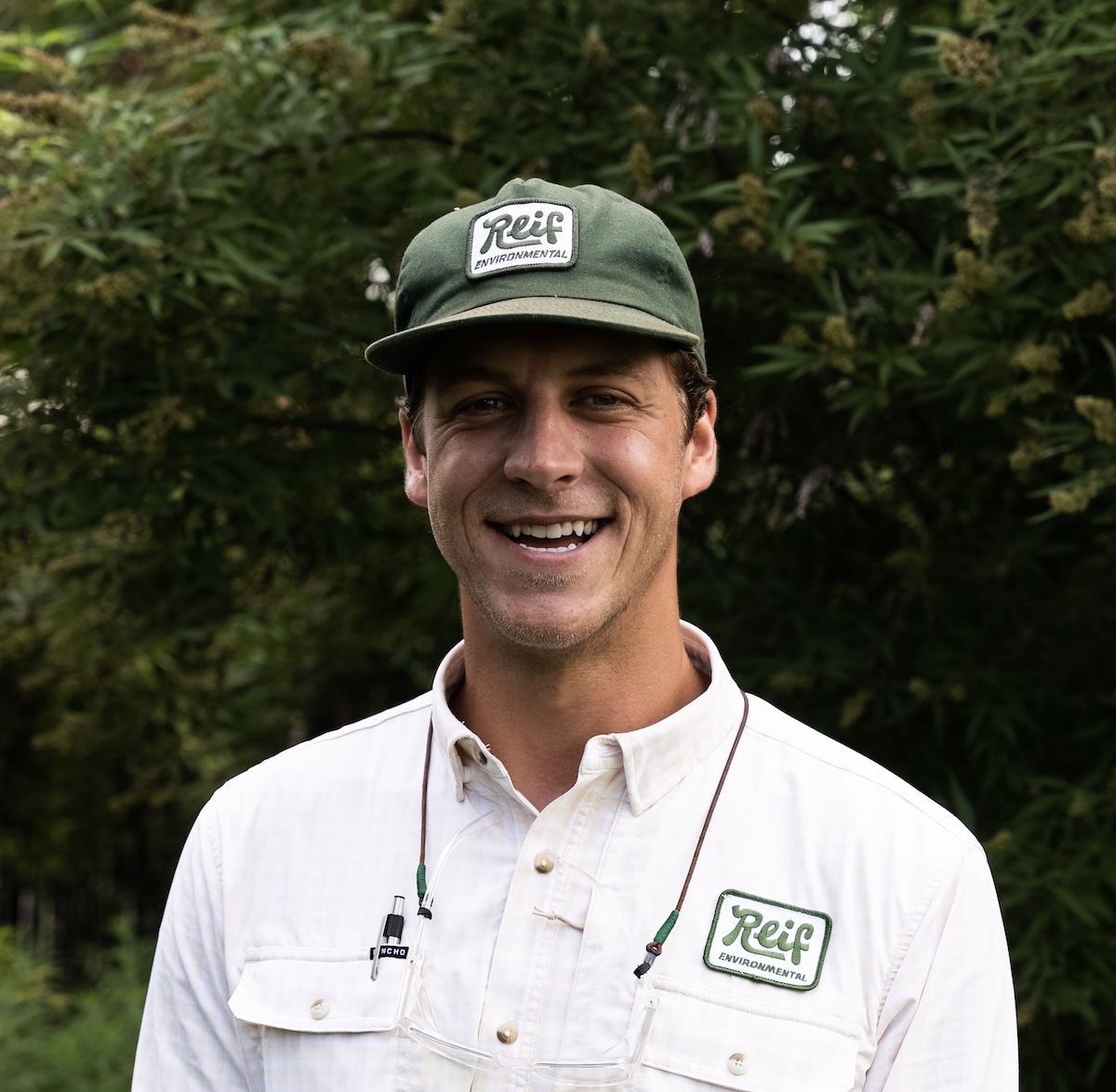 Smiling man wearing a green Reif Environmental cap and white shirt with Reif Environmental logo, standing outdoors with green foliage background.