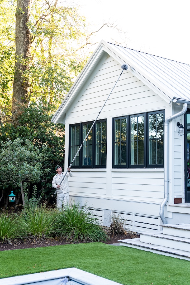 Person in white outfit cleaning high exterior window of white house using a long pole brush.