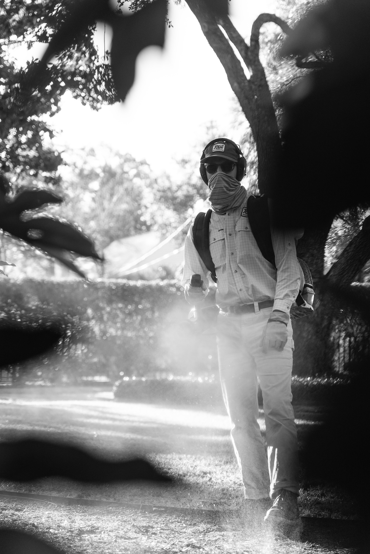 Person wearing protective gear, including headphones, sunglasses, and face covering, using a backpack sprayer in a garden.