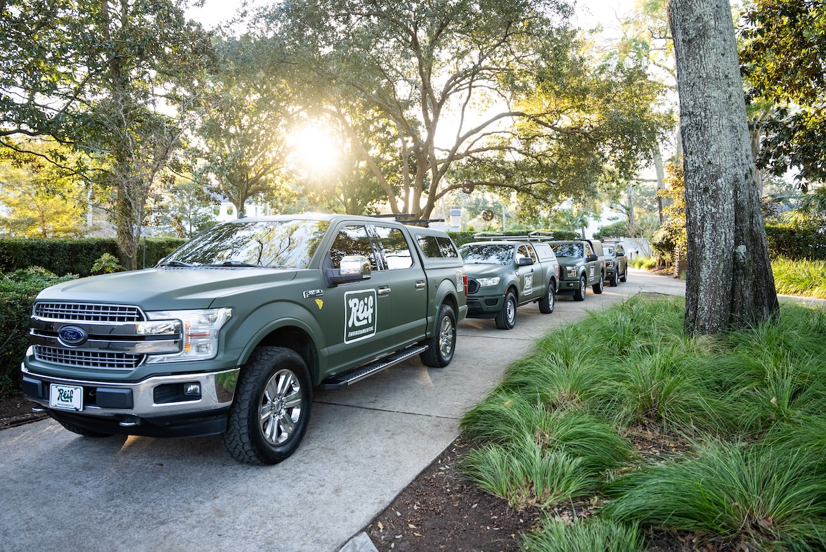 Line of green Reif Environmental pickup trucks parked on a sunlit driveway surrounded by trees and greenery.