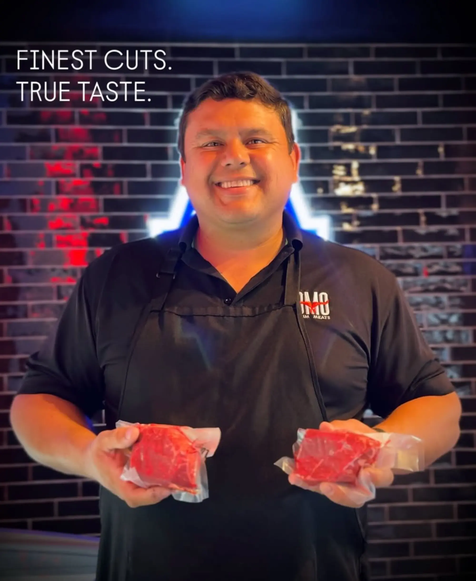 Smiling man in black apron holding two vacuum-sealed cuts of raw meat in front of a dark tiled wall with the text 'FINEST CUTS. TRUE TASTE.'