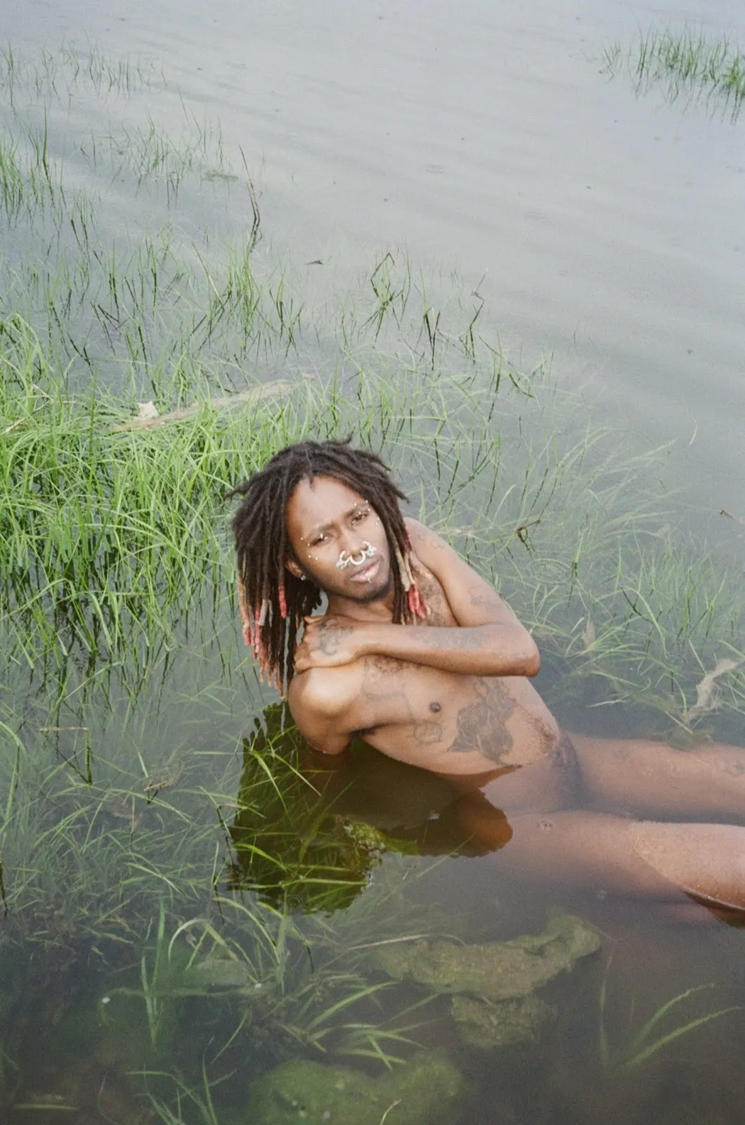 African American man sitting in grass-filled pond naked, wearing nose rings with facial piercings, fallen angel by morrison gong.