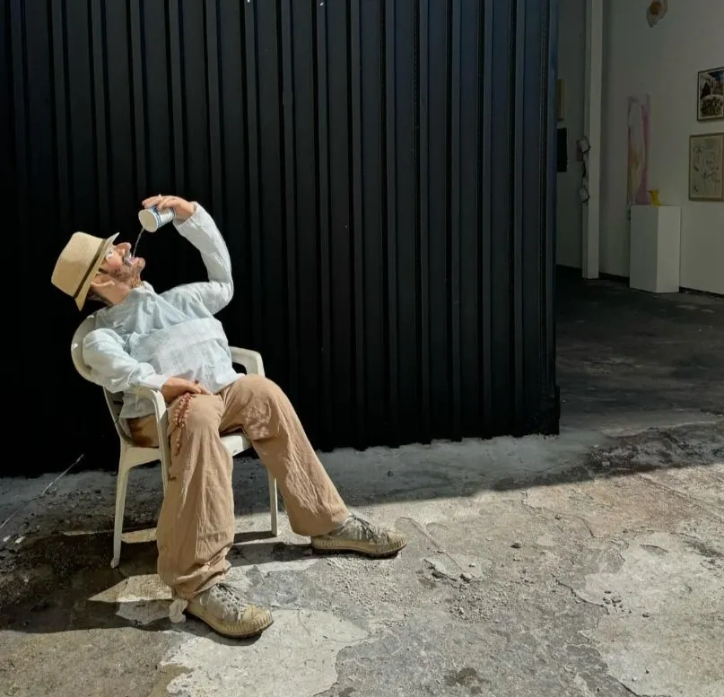Male Mannequin with blue collared shirt, holding a sack and water cup, khaki pants and straw hat drinking water on chair in front of gallery. Chris Retsina Taverna Man, Artists and recipes Marvin Gardens.