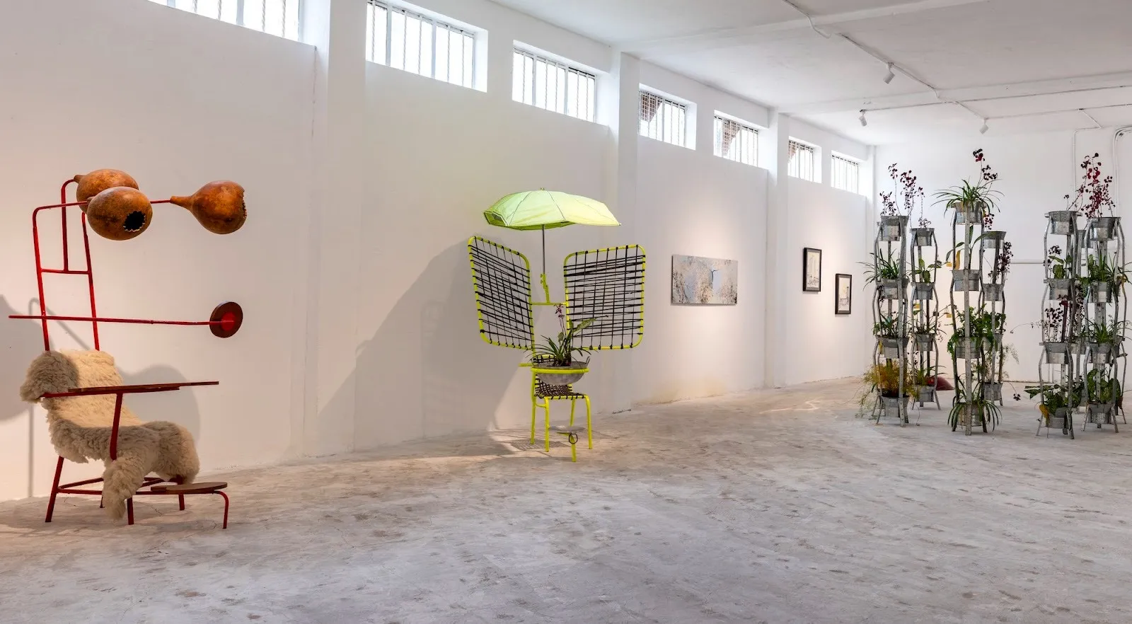 Installation view of Hakuna Futura gallery exhibition, two chairs with colorful welded metal extensions and shade with plants, paintings, drawings, and prints, Kairos Futura.