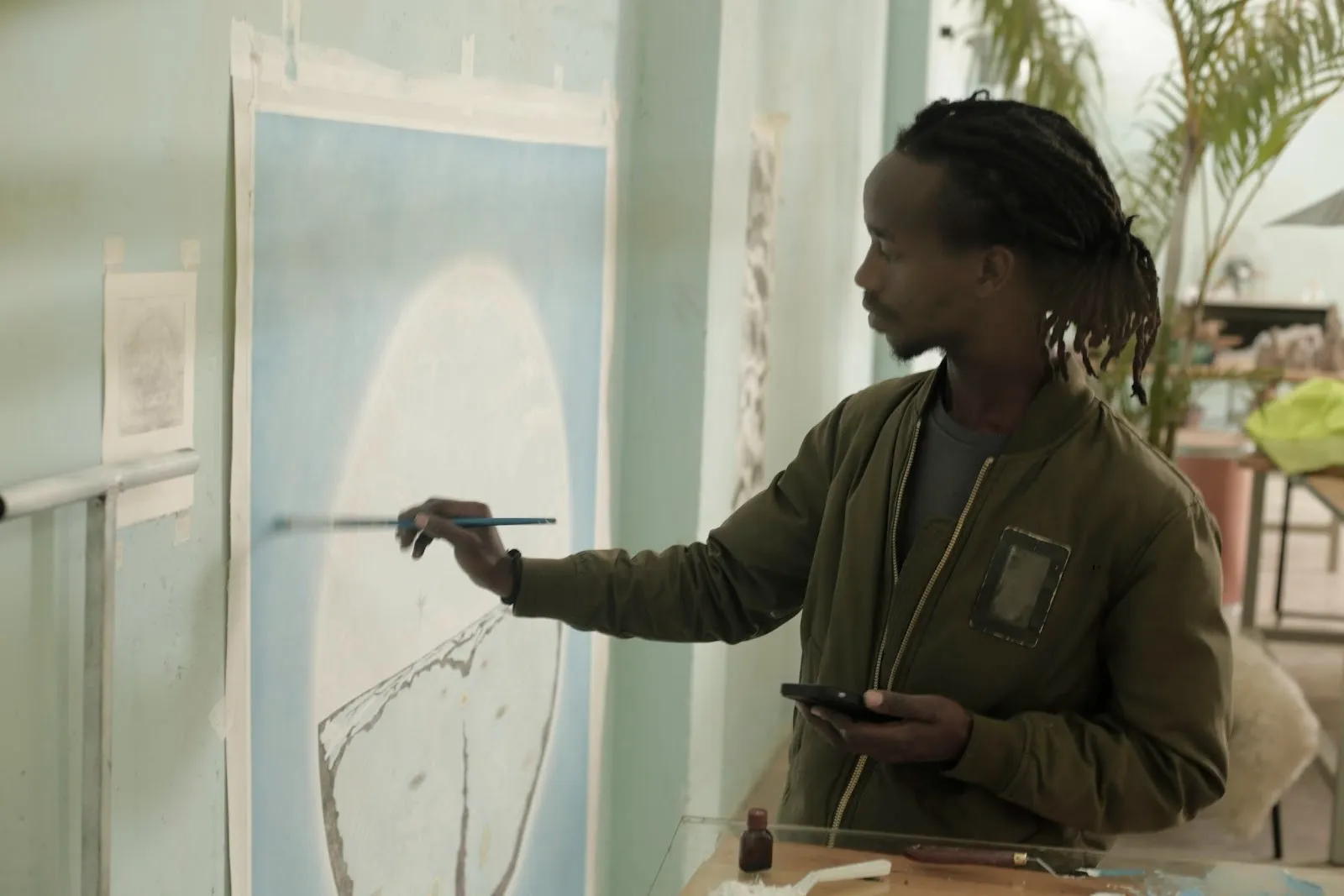 Man with Afro braids and bomber jacket holding a phone for reference photo while painting the Utopia within, Lincoln Mwangi from Kairos Futura.
