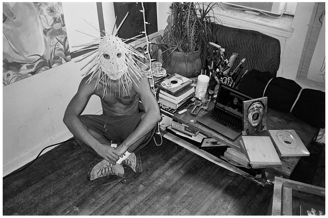 Guatemalan American artist Gio Black Peter sitting topless on the floor with spiky mask, next to a pile of CDs and books in Chinatown studio living space, queer artist, black and white portrait by Waltpaper.