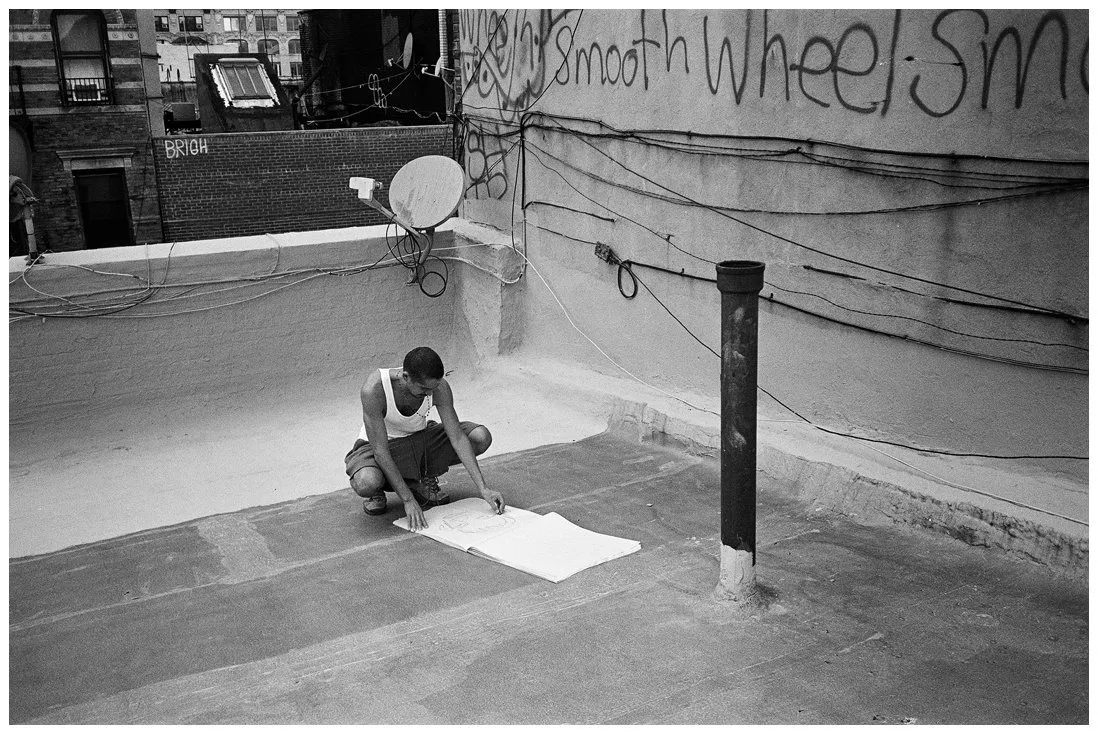Guatemala and American artist Gio Black Peter squatting on Chinatown rooftop in front of drawing pad, graffiti and wires in the background, queer artist, black and white portrait by Waltpaper.