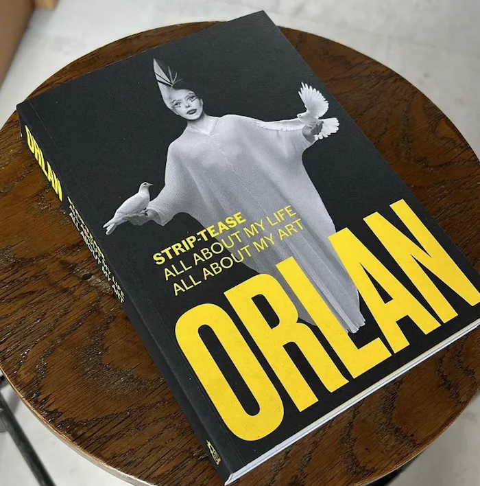 Book with black-and-white photograph of a woman in cape holding two pigeons on the cover on top of a desk, French carnal artist ORLAN's autobiography Striptease.
