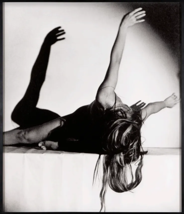 Black and white photograph of woman dancer holding arms up and head down with flowing long hair, sitting on a white platform casting sculpture-like shadow, ORLAN corps-sculpture.