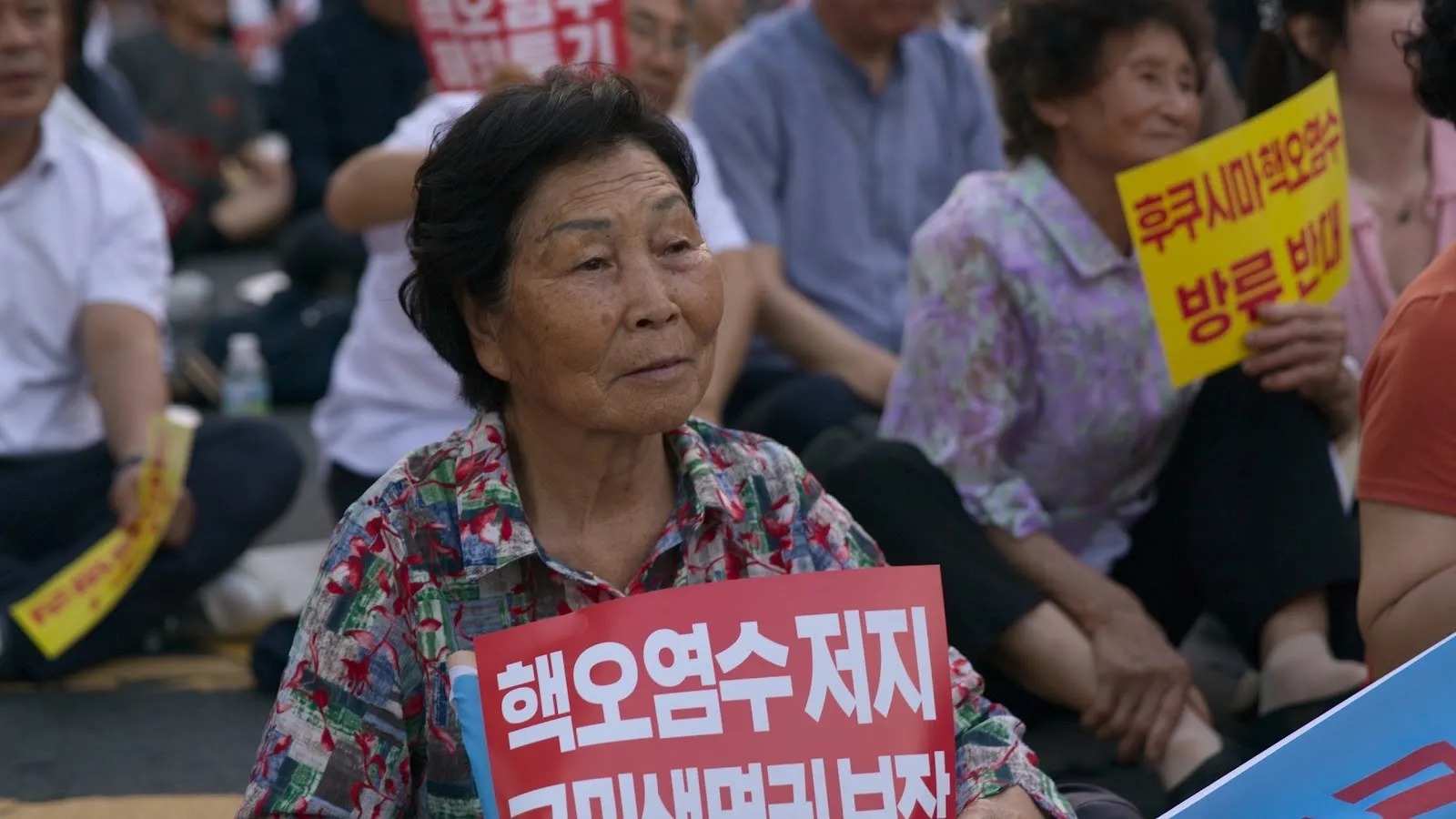 Korean Jeju Island female divers sitting on the ground holding signs to protest against neoliberal government policies, advocating for climate justice, photo still from the last of the sea women by Sue Kim.