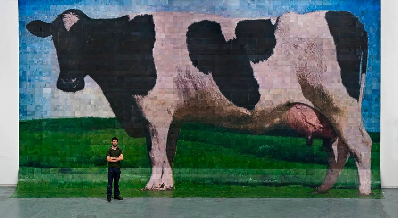 Male artist wearing black outfit standing in front of huge mural of a cow sending on grassland, Petros Lales paint on cardboard, collaged digital aesthetic pixel wall.