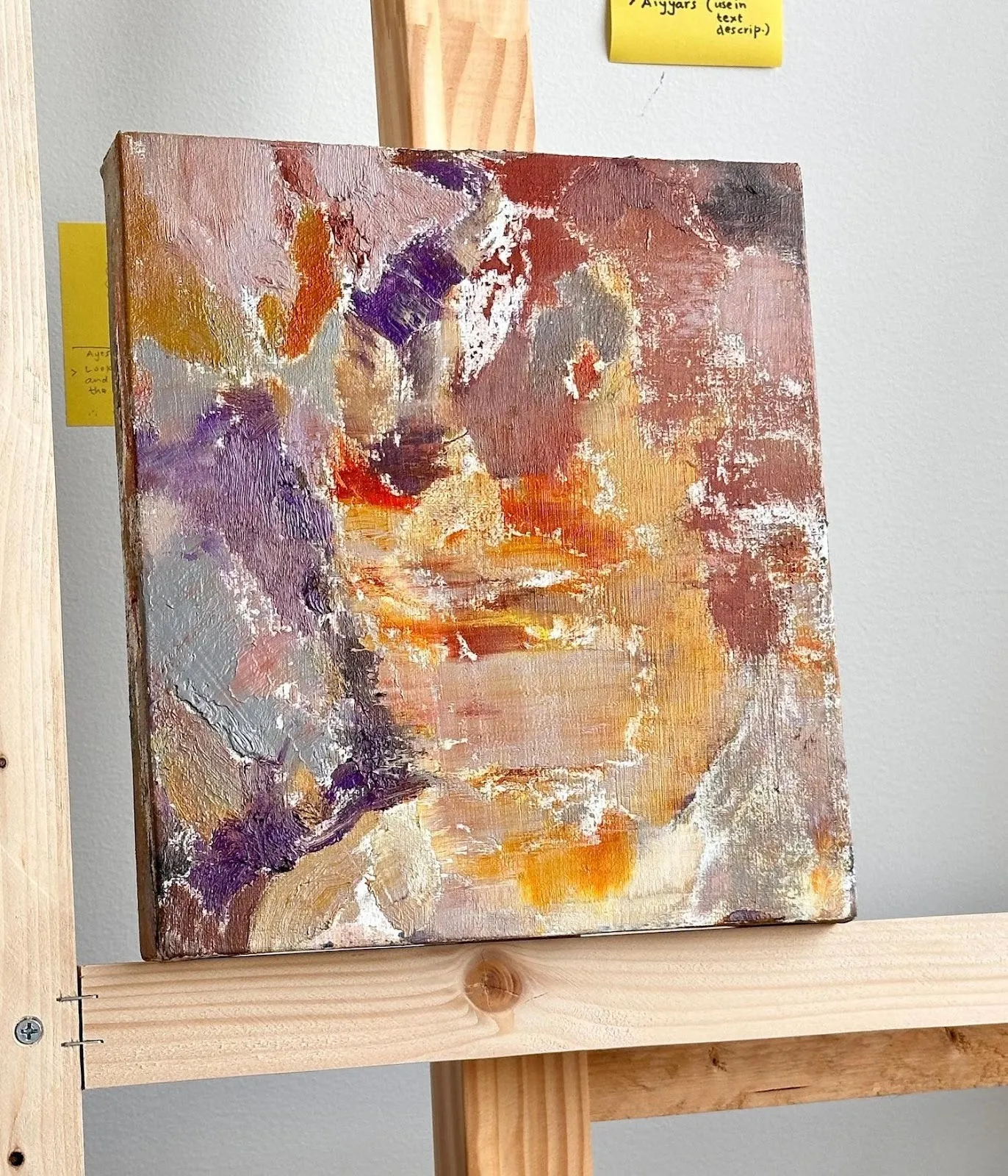 Oil painting with orange yellow red and purple paint sitting on easel against white wall with sticky notes, Studio photo of Indian figurative artist Kuldeep Singh.